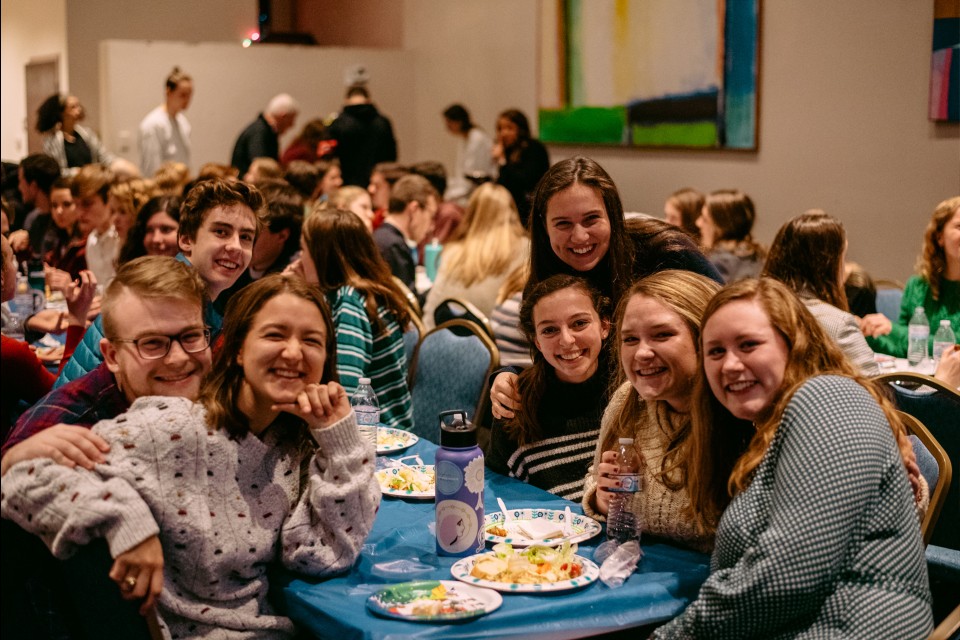 Students smile at the camera while sitting at a long dining table with plates of food.