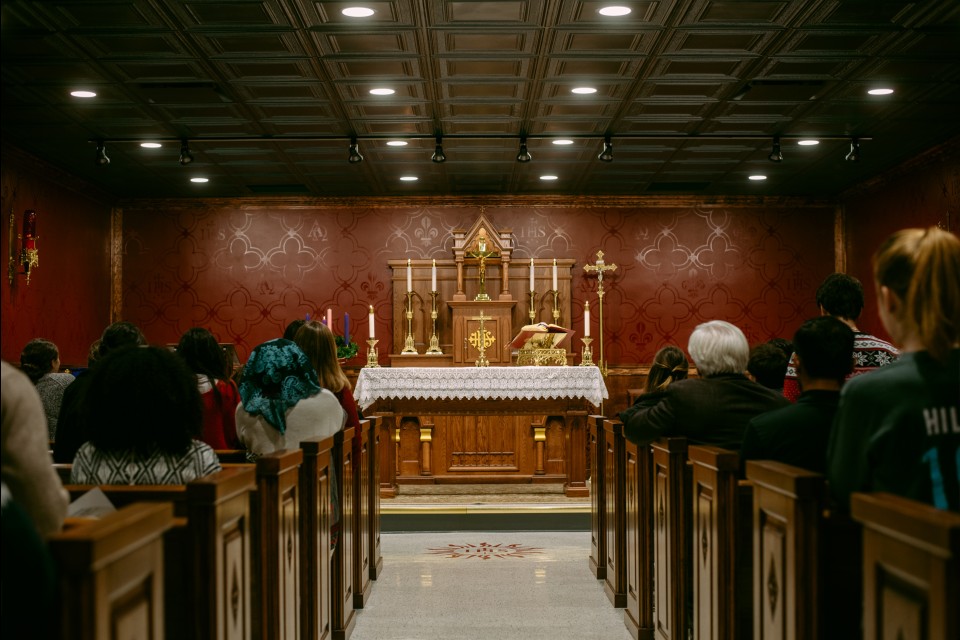 People sit, seen from behind, in the Catholic Studies Centre Chapel, looking at the altar. The altar is made of wood with a table covered in a lace cloth and candles, crosses and other accessories in gold.