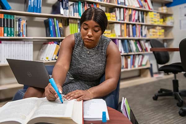 A woman sits at a table in the Academic Resource Center where she highlights from her notes. A shelf of resources, including books, is behind her.
