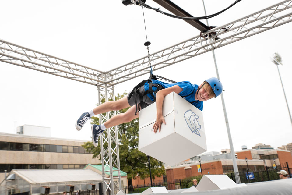 An attendee of the 2023 Homecoming and Family Weekend Pre-Game Celebration hands off a prize won as part of the human claw machine game while dangling from a harness. Photo by Luke Yamnitz.