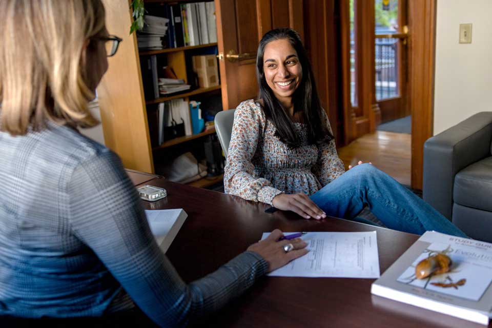 Image of a student sitting talking with a professor at their desk.