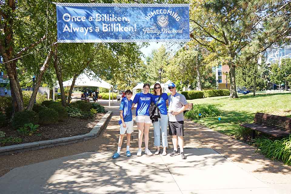 A family of four pose for a photo under a banner on campus that says once a Billiken always a Billiken Homecoming and Family Weekend.