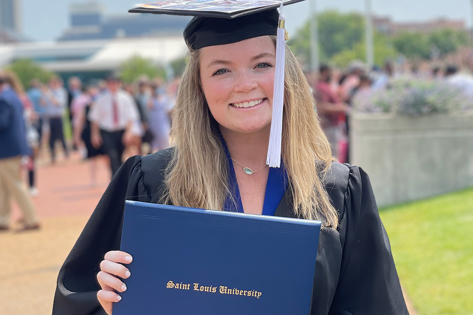 Becca Cary A woman wears a graduation cap and gown and holds a diploma outside on campus.