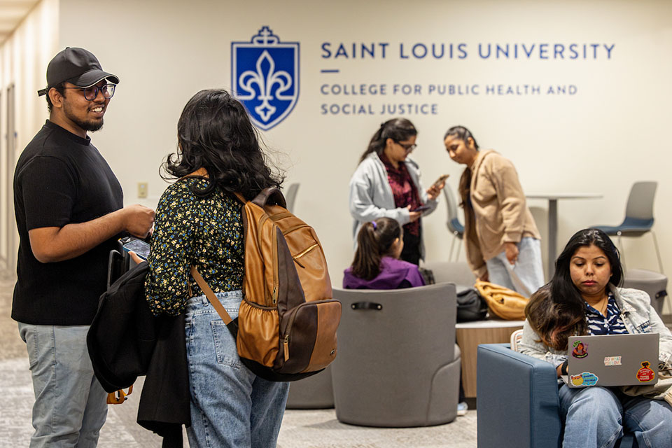 CPHSJ Students gather in a room on campus. The wall has a sign that says Saint Louis University College for Public Health and Social Justice.