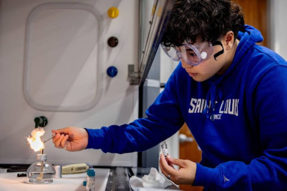 A student wearing safety goggles works in a lab with a burner that has a flame.