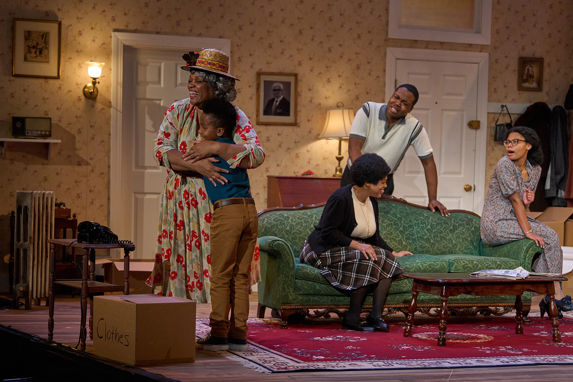 A grandma in a flowered dress and hat hugs her grandson as the rest of the family sits in the living room with varying expressions.