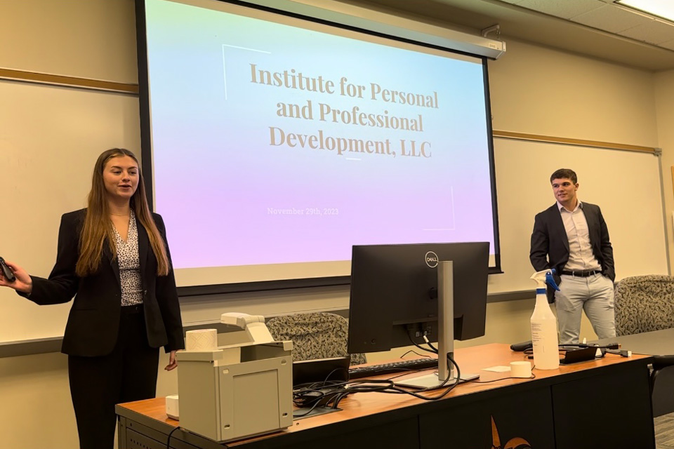 Two students wearing business clothes stand in front of a presentation screen that reads "Institute for Personal and Professional Development, L L C". 