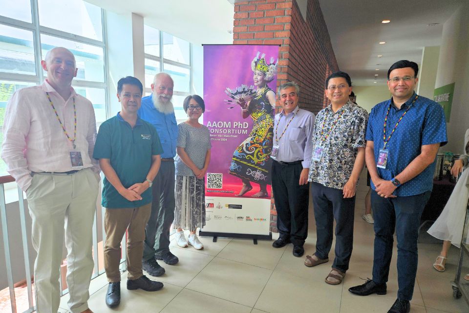 A group of conference attendees stand on either side of a banner that reads AAOM Ph.D. Consortium with an image of a Malaysian dancer in a colorful folk costume.