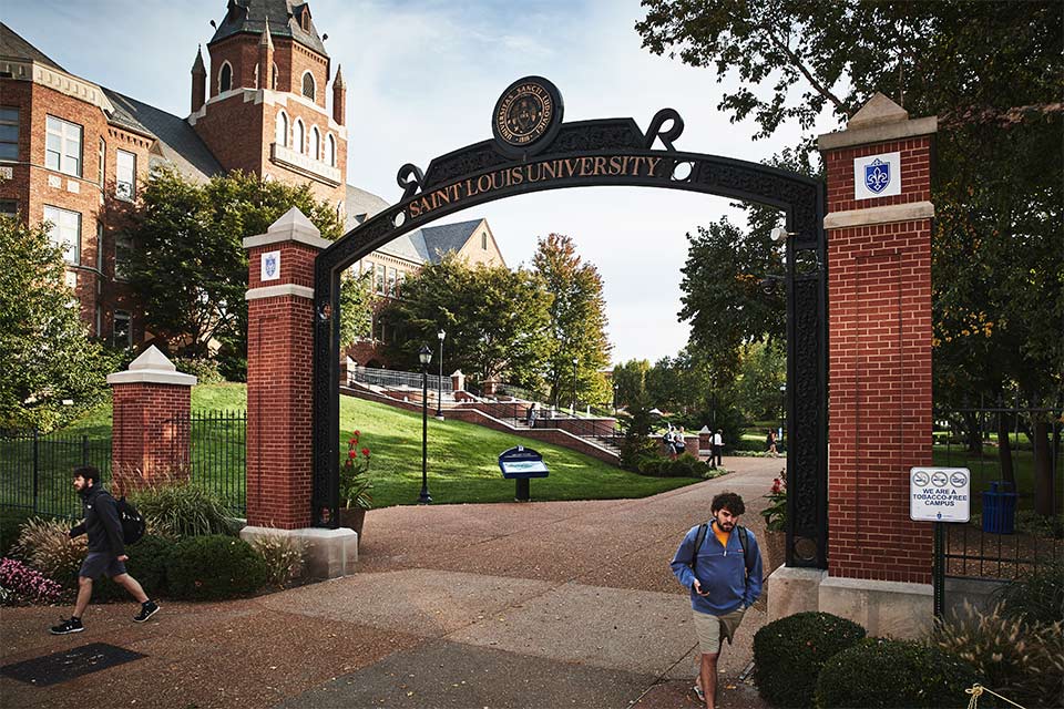 Students walk through the Saint Louis University archway at the entrance to campus with the Chaifetz School's Cook Hall in the background.