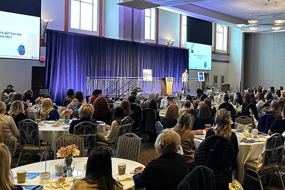 Attendees sit at tables looking at a speaker on a platform.