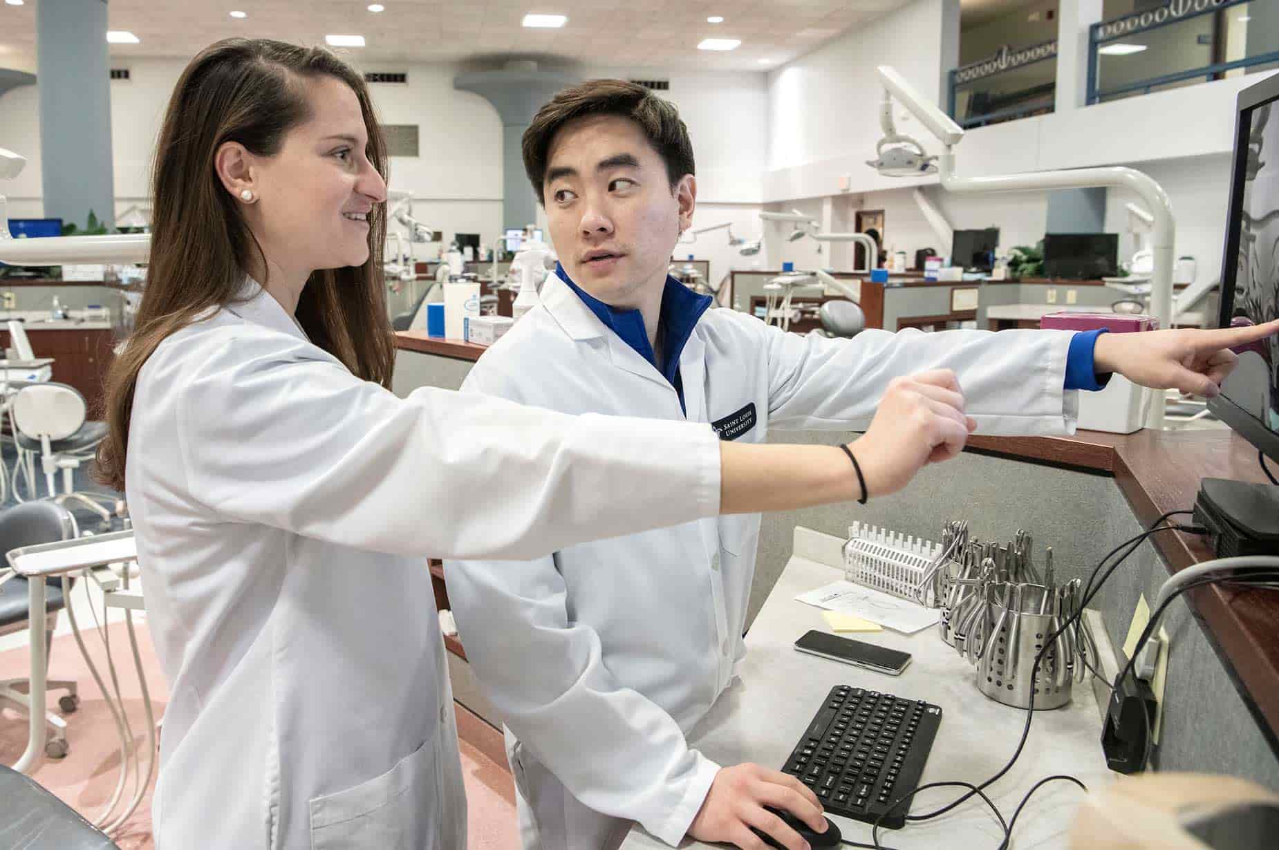Two CADE residents wearing white lab coats work at a computer workstation. The male resident points to something on the computer screen.