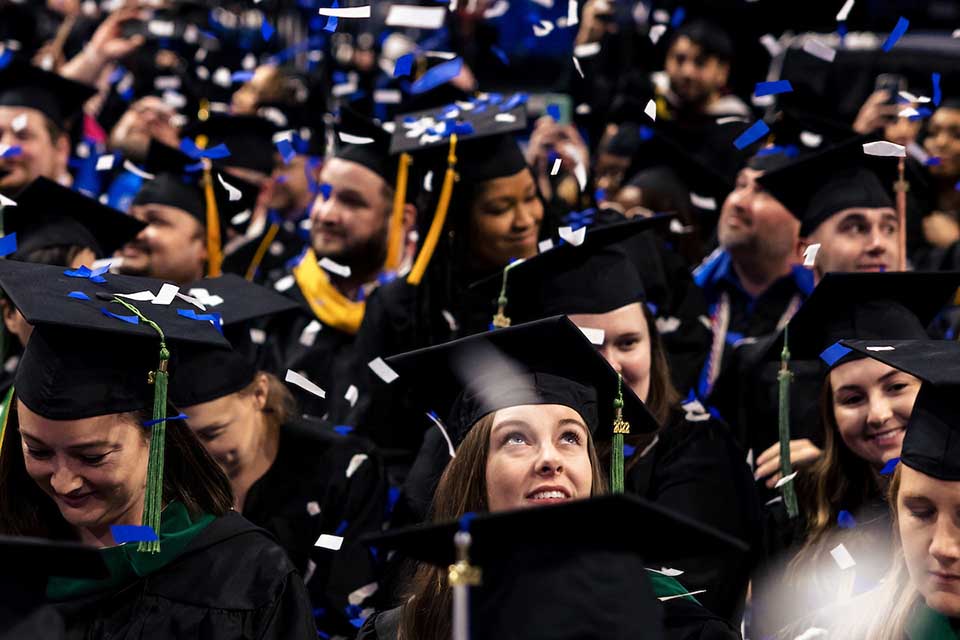 A student in cap and gown surrounded by a crowd of graduates looks up as confetti falls