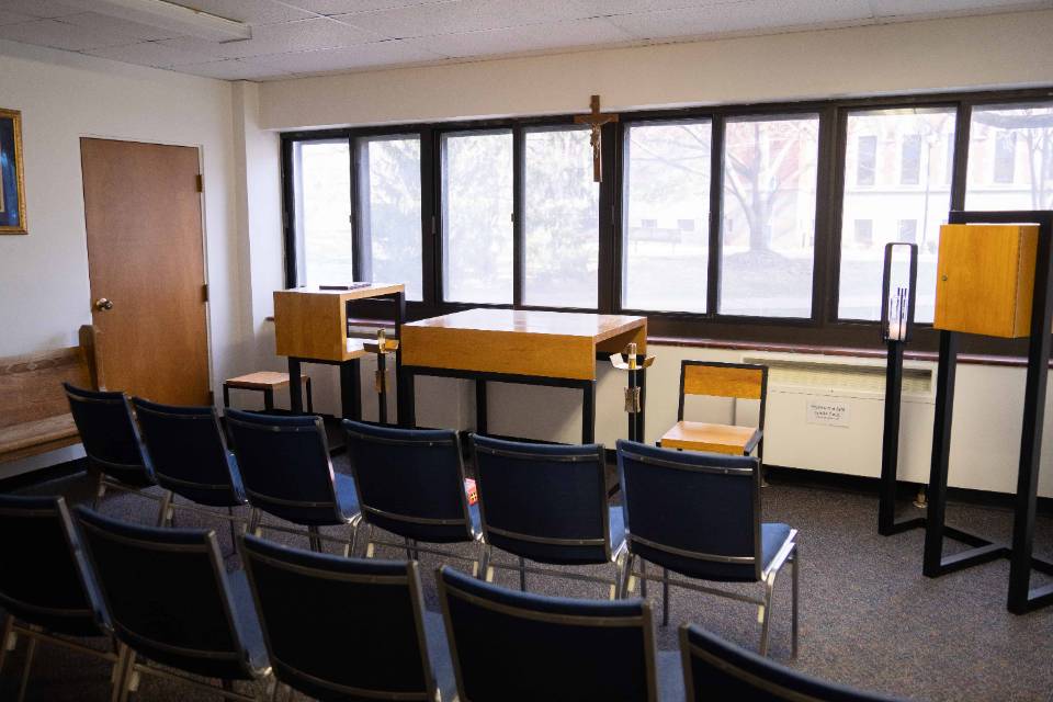 Chairs are seen from behind in two rows, facing a wooden altar table, chairs, crucifix and candle holder, along with a bank of windows.