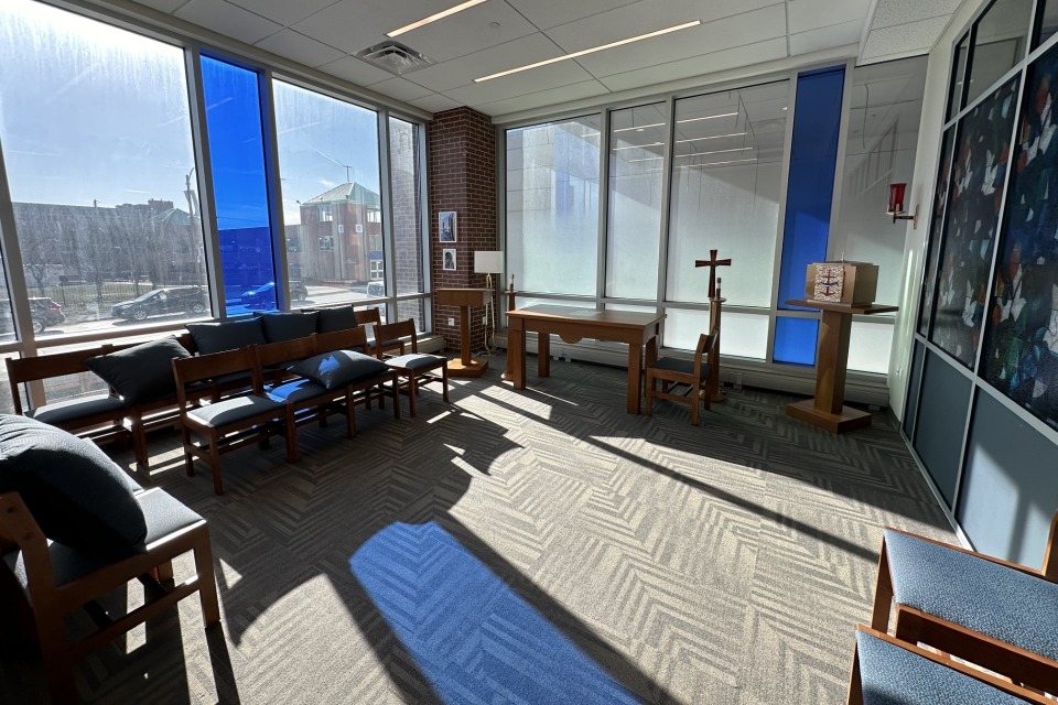Interior view of a room with padded chairs lining the walls, an altar table, cross, tabernacle and candle holders. There are large windows along along the wall with two stained blue. Sunlight is streaming in, and the sunlight is reflected on the carpet.