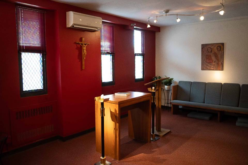 Interior view of a room with an altar, crucifix lectern and candle holder at the front and center, with a padded pew along one wall. A religious painting is visible.