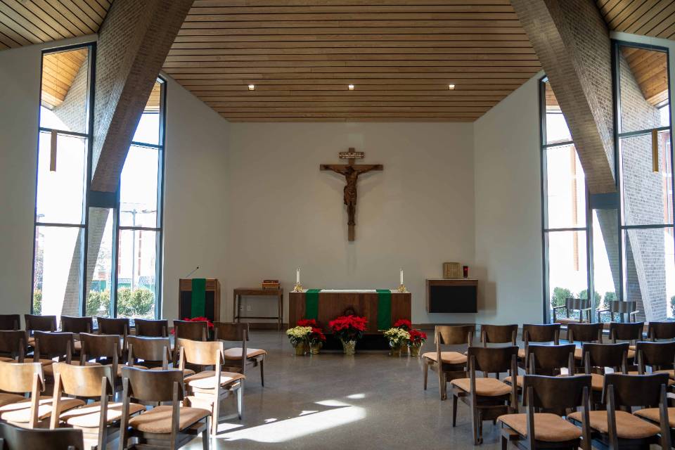 View of a chapel with rows of chairs seen from the back, with an aisle down the middle. At the front, there is an altar, crucifix. Red and white poinsettias are on display. There are tall windows on each side, with a slatted wooden ceiling.