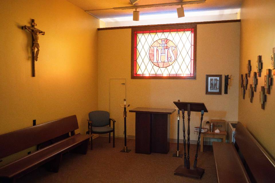 A room with wooden church pews on each wall and an altar area in the front and center. A crucifix and framed art is on the wall. Behind the altar is a stained glass window that reads I H S.