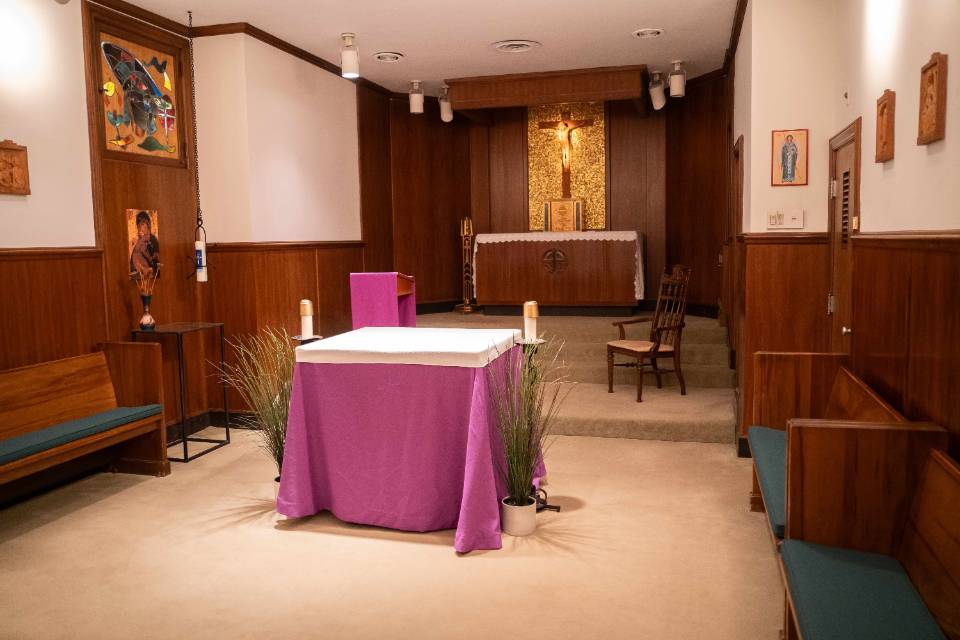 A room with church pews along each wall. There's an altar table in the center. In the background, behind the altar table, there is a long cabinet with a tabernacle sitting on it, with a crucifix on the wall, surrounded by gold wall decoration.. Framed religious artworks are visible on the walls.