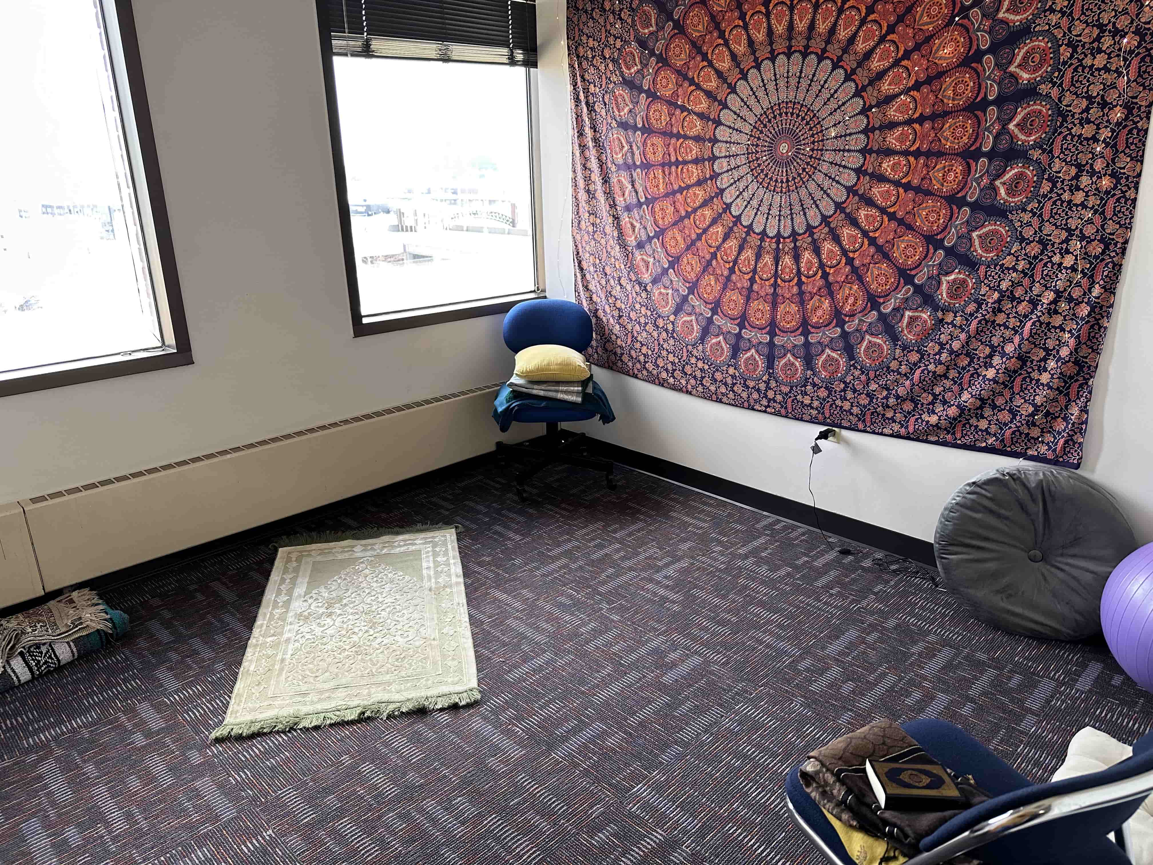 A room has a prayer rug on the floor as well as a mandala tapestry on one wall. Chairs and cushions are also visible.