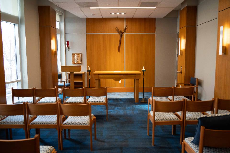 Chairs are seen in rows from behind with an altar at the front of the room. An abstract, modernist crucifix is on the wall.
