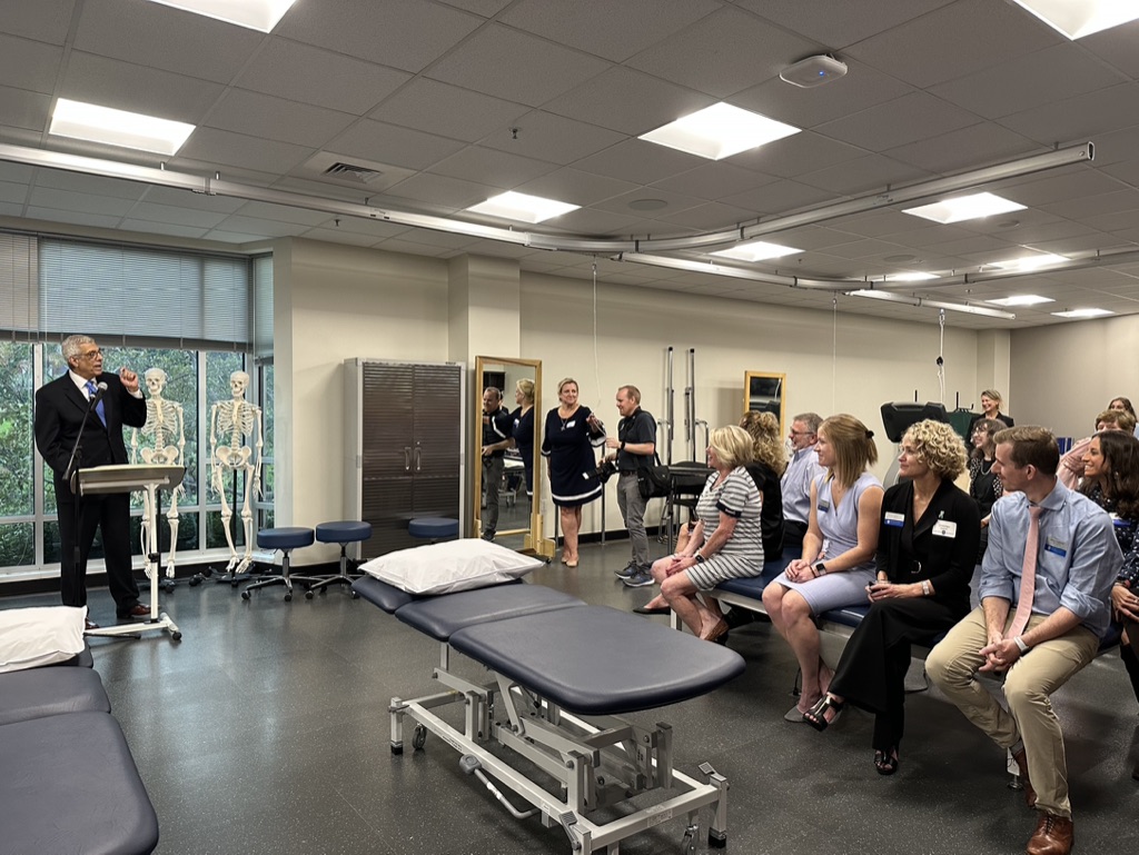 Audience members sit in chairs while president Fred Pestello speaks at a lectern. The room contains items such as a physical therapy table, display skeletons and other tools.