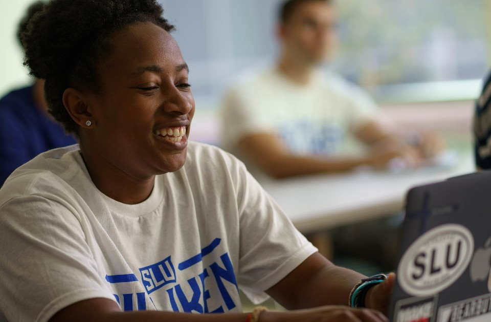 A student wearing a SLU T shirt smiles while working on a laptop computer in a classroom.