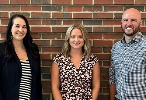 The PRiME leadership team stands in front of a red brick wall and smiles. Left to right are Associate Director of Operations Ashley Donaldson Burle, in a black jacket and black and white striped shirt;  Post-doctoral Research Associate Misti Jeffers, Ph.D., in a red blouse with white flowers, and Director of Research and Evaluation Evan Rhinesmith, Ph.D., in a blue-checked button-down shirt.