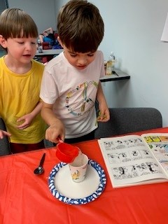 Two campers pour ingredients into a paper cup while standing next to a table during an activity.