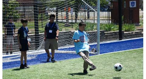A student kicks a soccer ball on a field while two others, standing in a goal, watch.