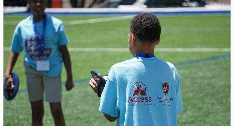 A student wearing an Acess Academies T-shirt is seen from behind while talking to another student on a grassy field.