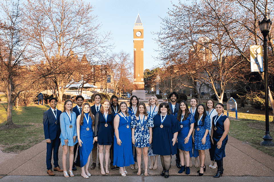 A group of 18 students wear medals around their neck with the clocktower over their heads in the background.