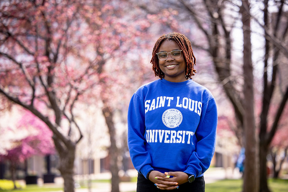 Malisyah Vann poses outdoors on the SLU campus with red locust trees in the background and smiles.