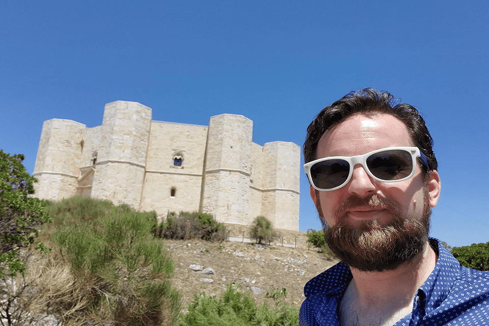 A man wearing sunglasses looks at the camera, with a stone castle in the background.