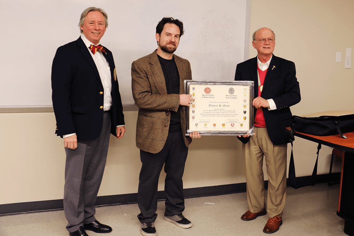 Thomas Morin stands with two representatives from the Military Order of the Crusades in a classroom holding a framed certificate. 