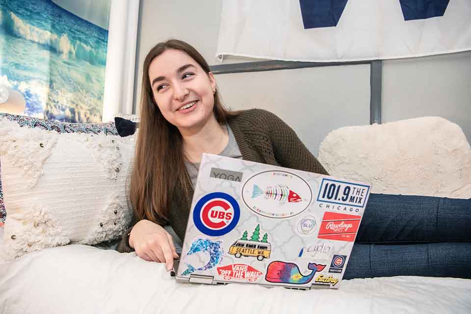A resident reclines on her side on a bed with pillows, she's smiling at something off to the side. A laptop with many stickers is in front of her.