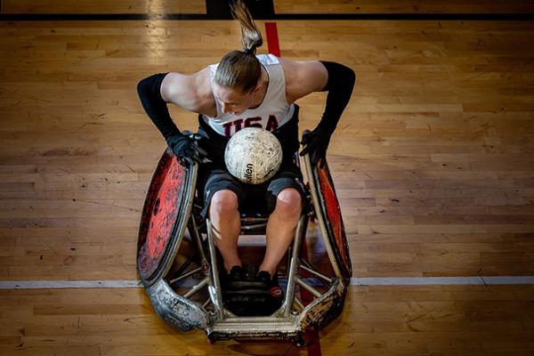 A woman with a ponytail sits in a wheelchair. A rugby ball is on her lap while she works the wheels on her chair. Her muscles are flexed with exertion.