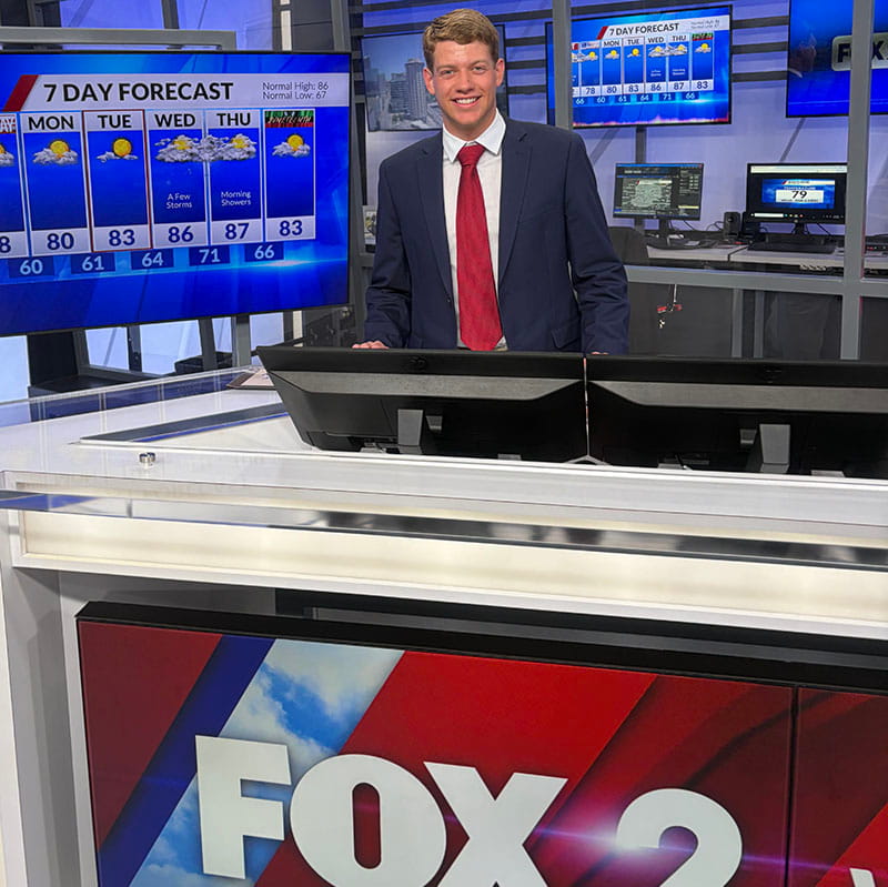 A student wearing a suit and tie stands on a television news set in front of a screen that says 7 Day Forecast