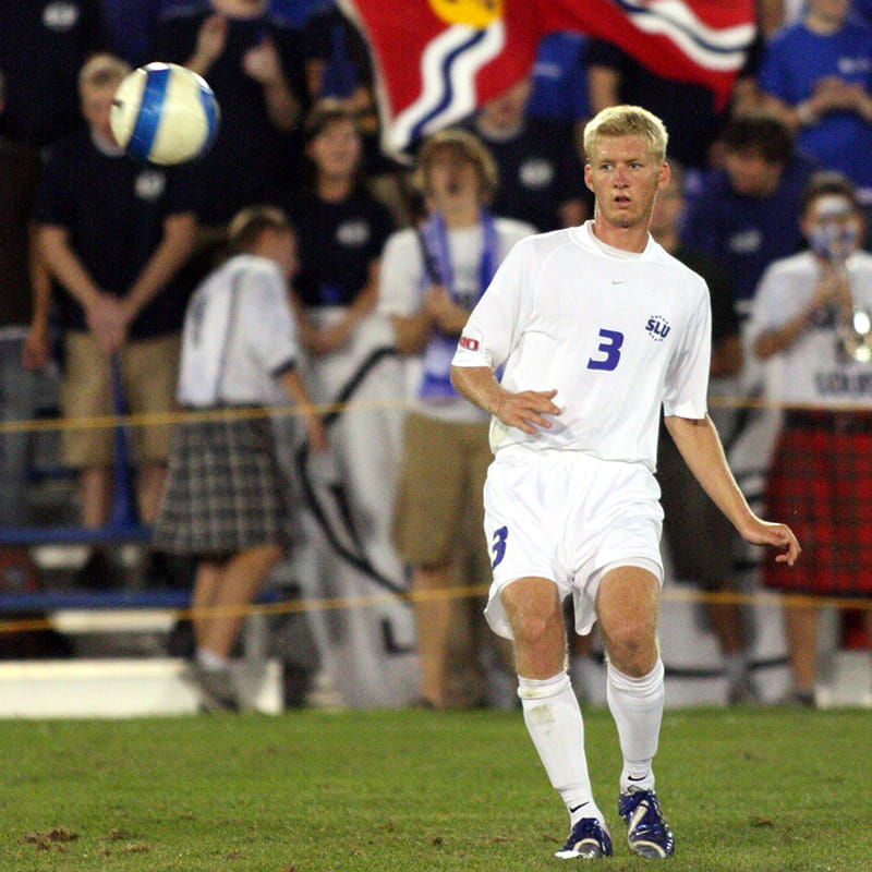 A soccer player wearing a SLU jersey number 3 eyes a soccer ball in the air.