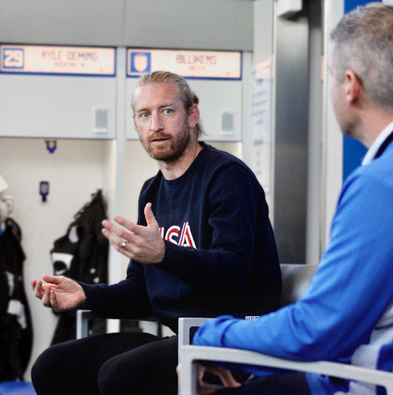 A man wearing a sweatshirt that says USA sits in a chair during a discussion.
