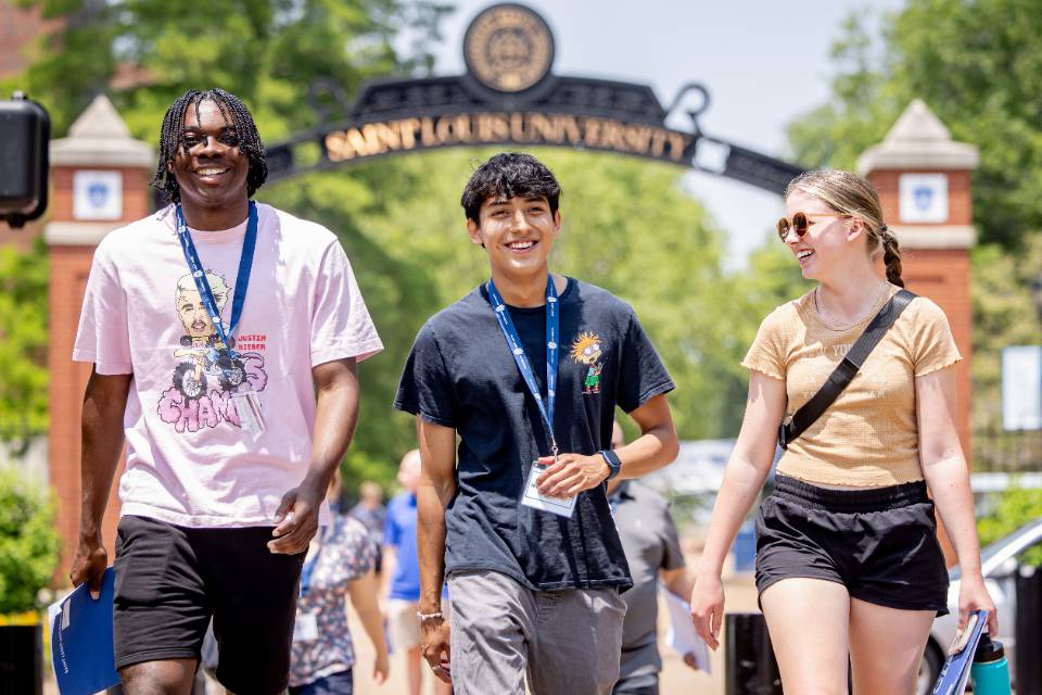 Three students with nametags on walk on the SLU campus with the arch saying Saint Louis University in the background.