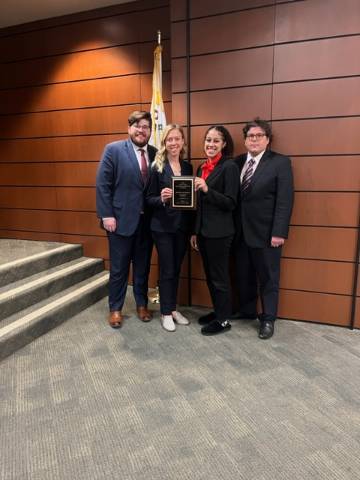 Four students wearing business suits stand against a wall holding a plaque