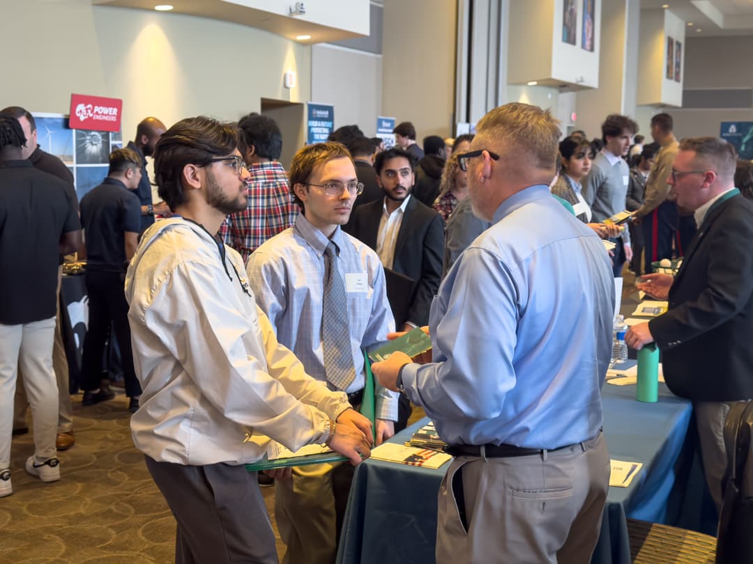 Two students talk to a prospective employer in a room filled with booths and people.
