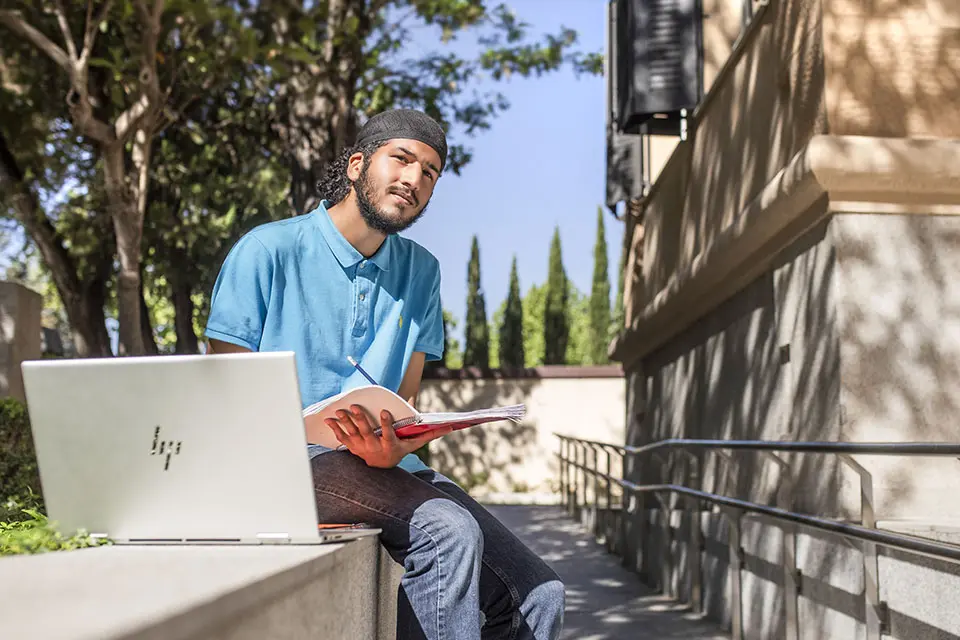 A student sitting on a stone ledge with a notebook in his hand and a laptop next to him.