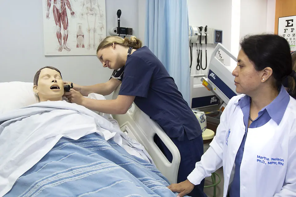 A student wearing medical scrubs checks the ear of a patient model on a hospital bed while an instructor observes.