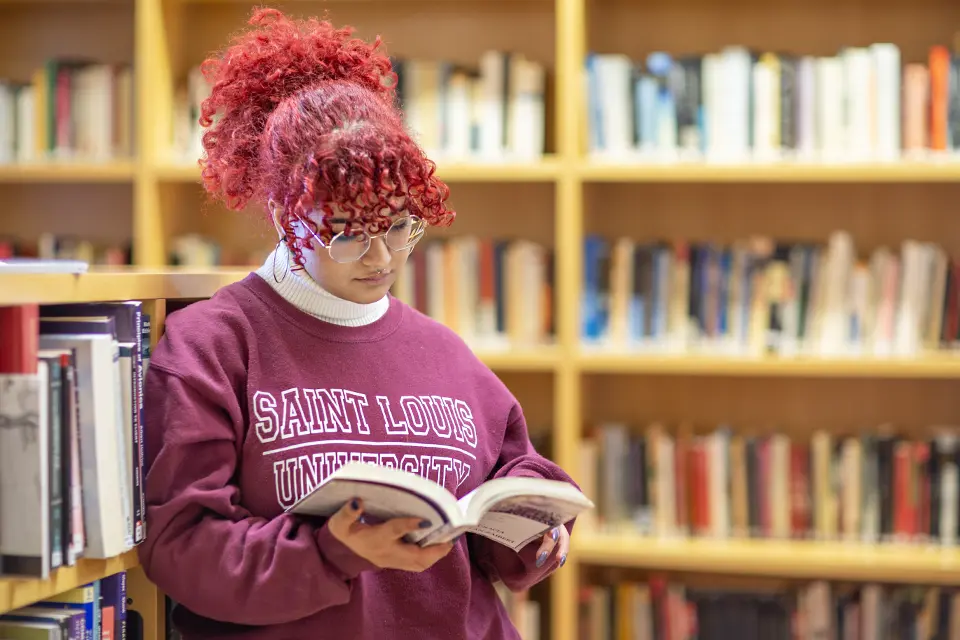 Student stands in front of book shelves, holding a textbook.