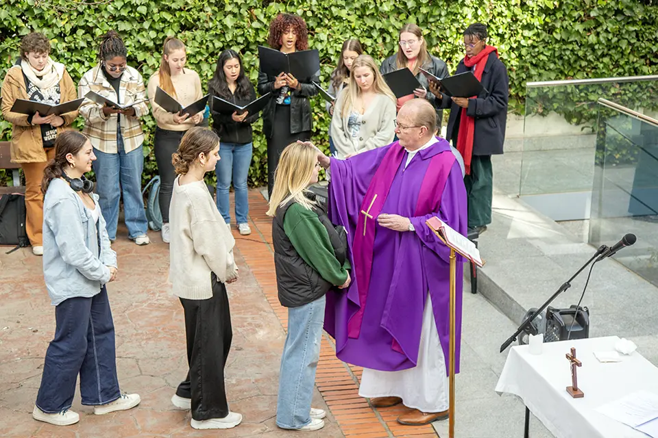 Students line up in an outdoor area while a priest marks a student's forehead. A choir of students stands nearby holding black choir books and singing.