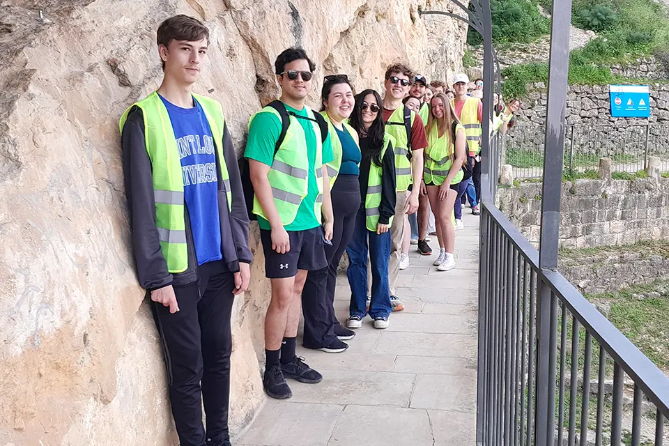 A group of students wearing reflective safety vests stands in a line on a walkway next to a rocky cliff side.