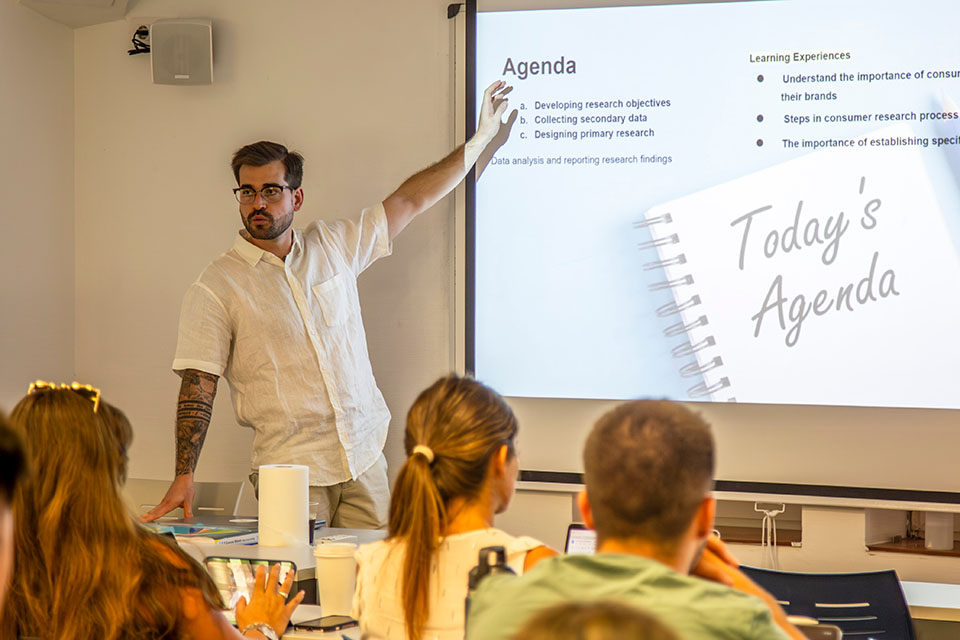 Miguel Garcia Miguel García faces a classroom of students while gesturing to a screen with a powerpoint slide that says Agenda.