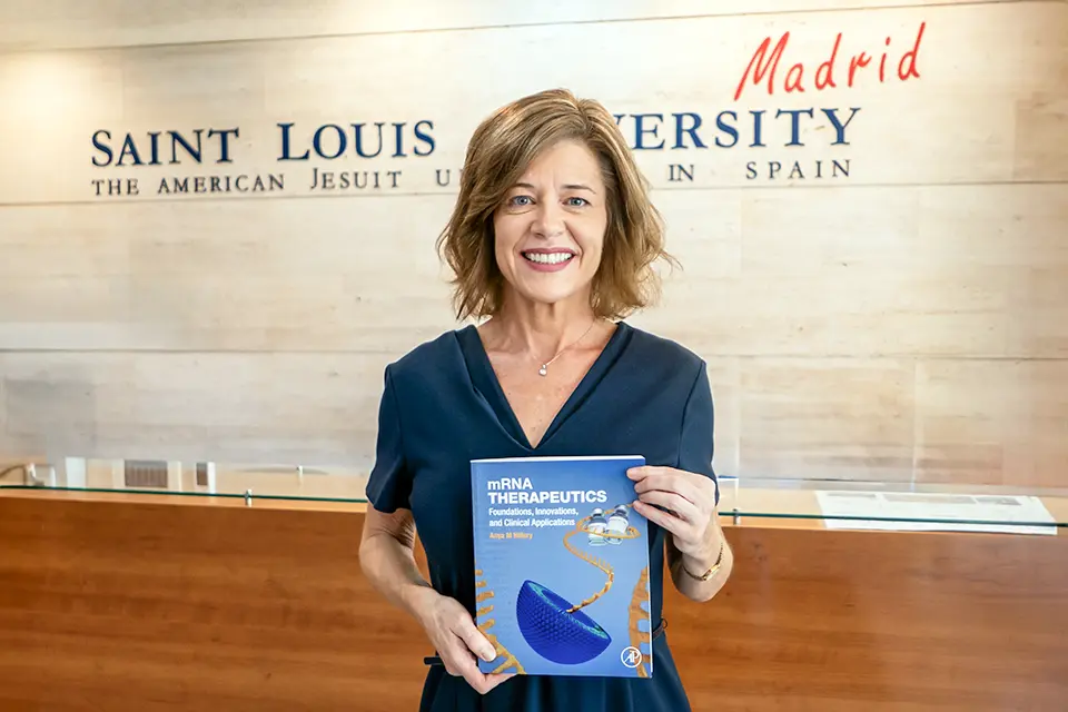 Woman holds book and poses for camera in front of reception desk with university logo on wall. Book title reads M R N A Therapeutics.