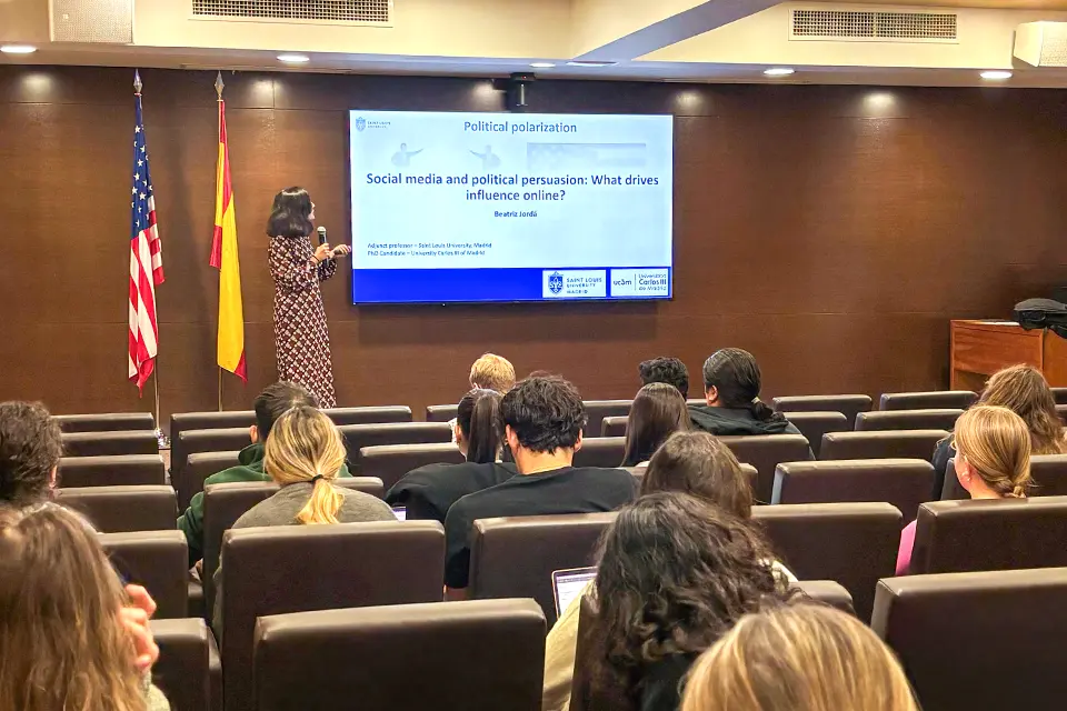 Woman with a microphone in hand looks at a slide of a presentation on a screen on stage in a small auditorium with students in the audience. The slide reads: Political polarization: Social media and political persuasion: What drives influence online?